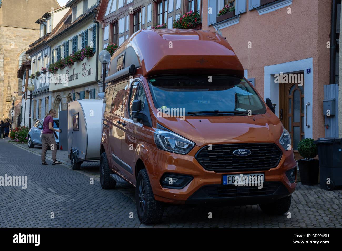 Une fourgonnette orange Ford transit Custom Nugget Westfalia avec un toit haut tirant une remorque en larme argentée sur une rue historique de la vieille ville. Banque D'Images