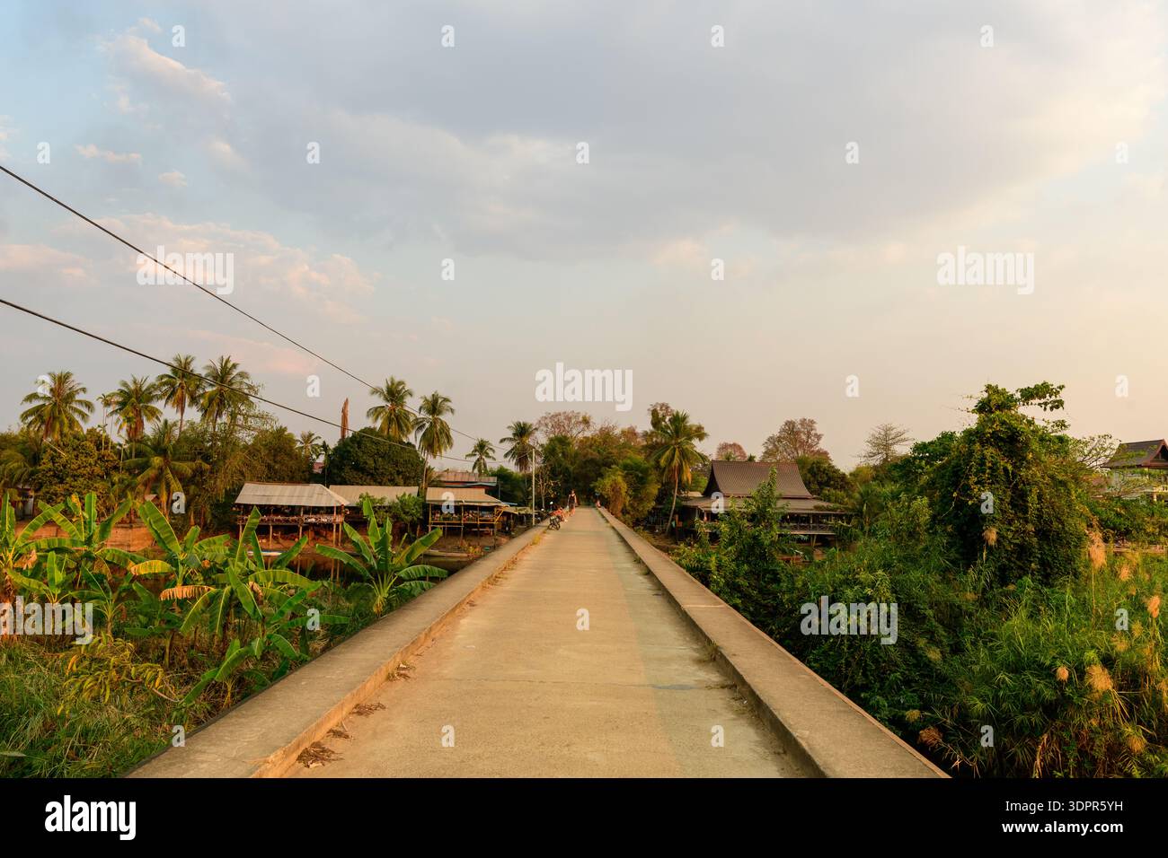 Lieu inconnu - 27 février 2020 : un pont en béton s'étend au loin avec une végétation tropicale, des palmiers et des maisons traditionnelles visibles des deux côtés sous un ciel partiellement nuageux. Banque D'Images