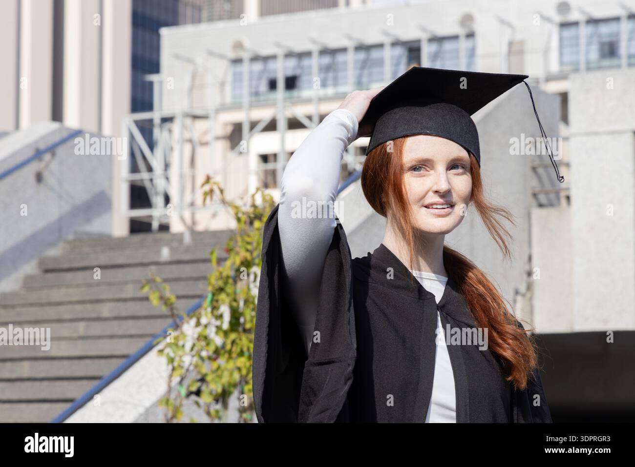 Femme diplômée debout sur des marches en béton sur le campus dans la casquette et la robe tenant mortarboard, espace de copie Banque D'Images