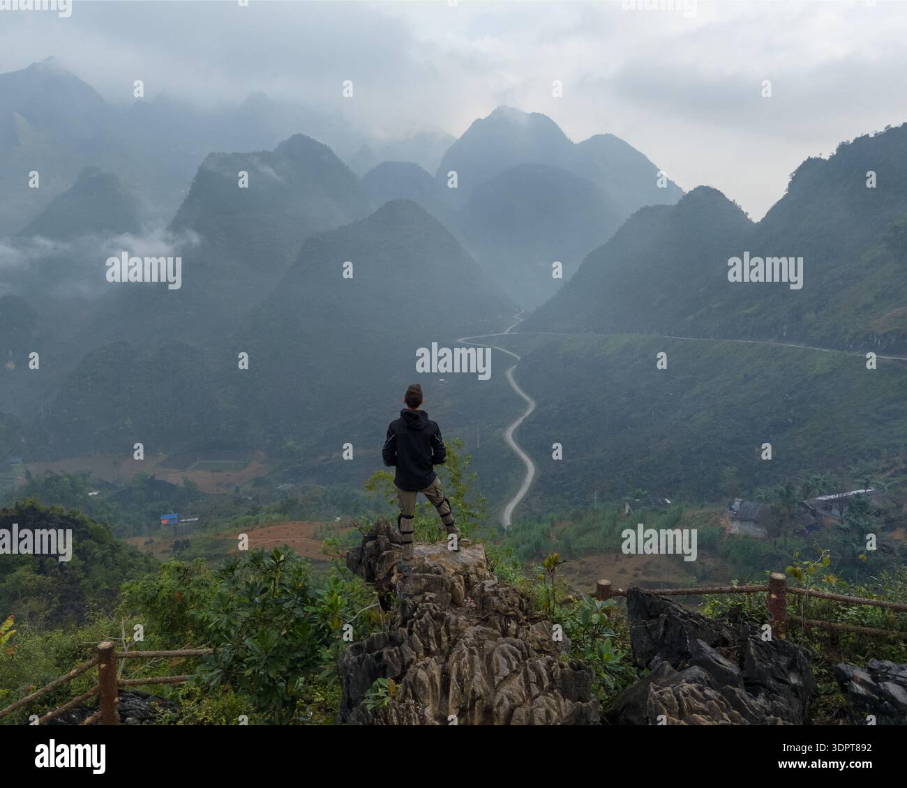 Une vue aérienne oblique montre une personne debout sur un point de vue de montagne rocheuse surplombant une route sinueuse et des collines karstiques boisées dans le nord du Vietnam. Banque D'Images