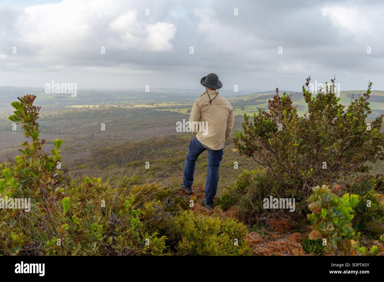 Une personne se tient sur une colline rocheuse regardant au-dessus du Bush vallonné et des terres agricoles lointaines sous un ciel nuageux. Banque D'Images