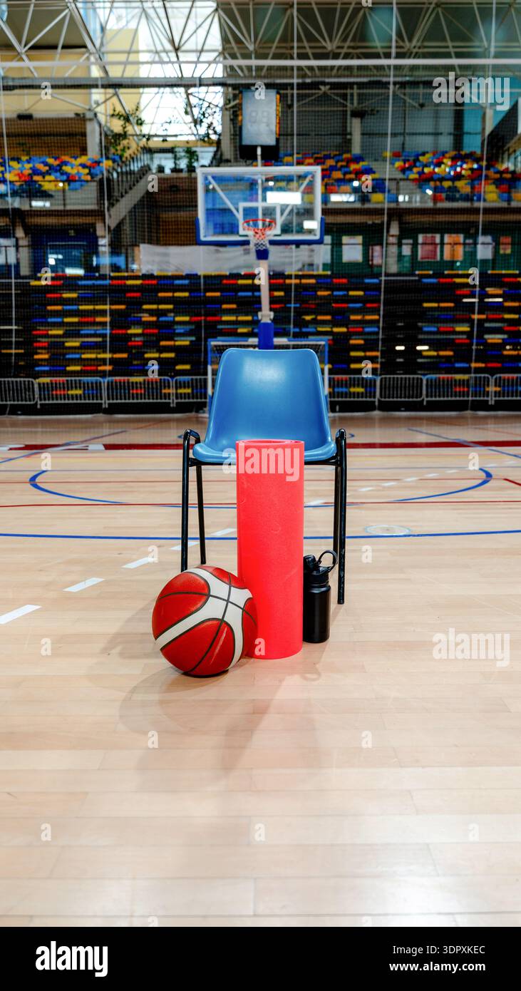 Une scène de terrain de basket-ball avec une chaise bleue avec un ballon de basket-ball, rouleau en mousse rouge, et bouteille d'eau noire à l'avant, mis contre des gradins colorés et Banque D'Images
