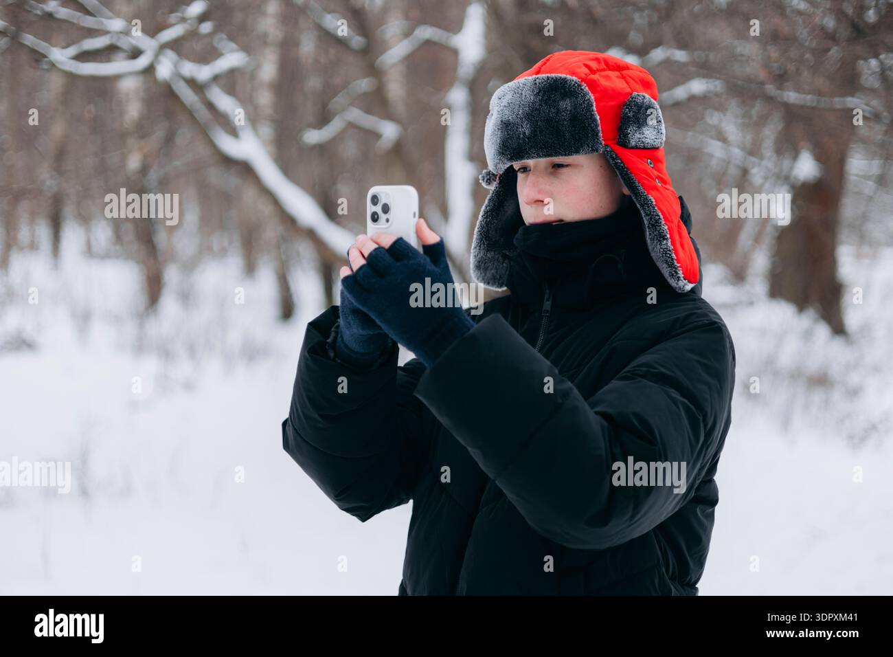 Un adolescent vêtu de vêtements chauds prend des photos avec son smartphone dans une forêt hivernale enneigée, capturant la beauté sereine de la nature pendant le Banque D'Images