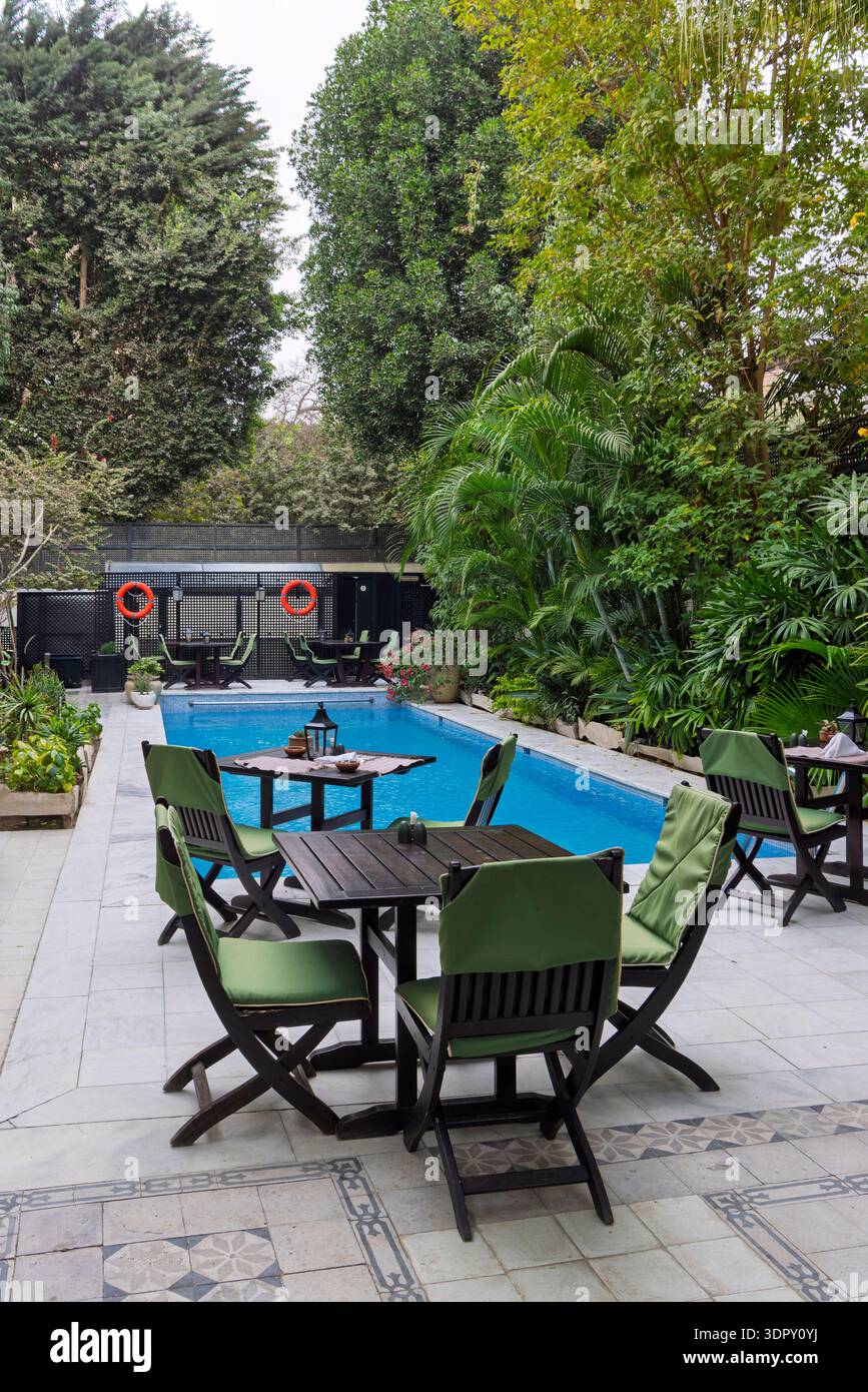 Un patio tranquille au bord de la piscine avec des chaises en bois rembourrées de vert et des tables à côté d'une piscine bleu clair, entouré de plantes tropicales denses et de grands tr Banque D'Images