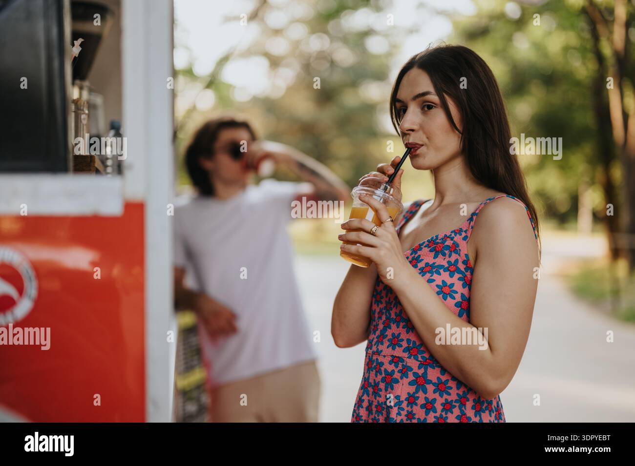 Femme profitant d'une boisson rafraîchissante de jus dans un parc ensoleillé à côté d'un camion de nourriture rouge Banque D'Images