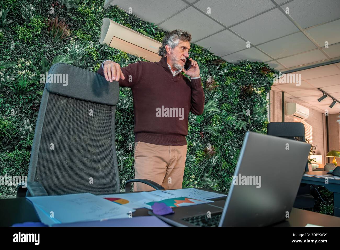 Professionnel senior discutant du travail sur smartphone tout en se tenant près d'un mur vert dans un bureau moderne avec ordinateur portable et documents Banque D'Images