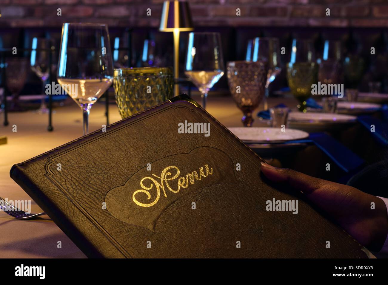 Gros plan sur la main de la personne tenant le menu relié en cuir tout en étant assis à la table du restaurant élégamment dressée. Ambiance gastronomique avec table, verrerie Banque D'Images
