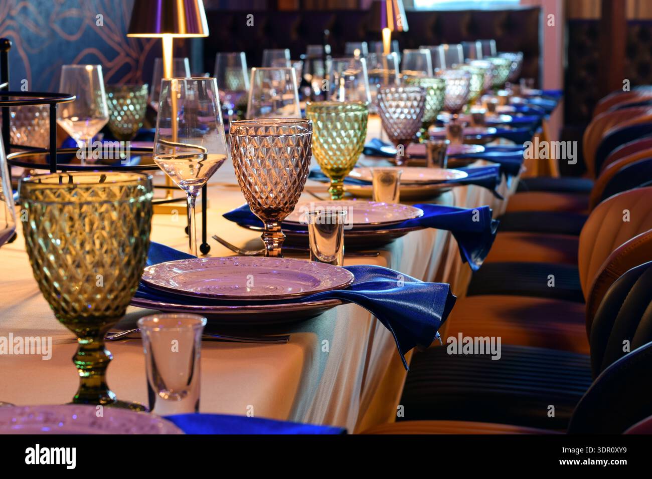 Élégante table de banquet ou de restaurant avec des gobelets colorés, des verres à vin, des serviettes bleues et des assiettes blanches. L'éclairage chaleureux crée une atmosphère de salle à manger formelle Banque D'Images