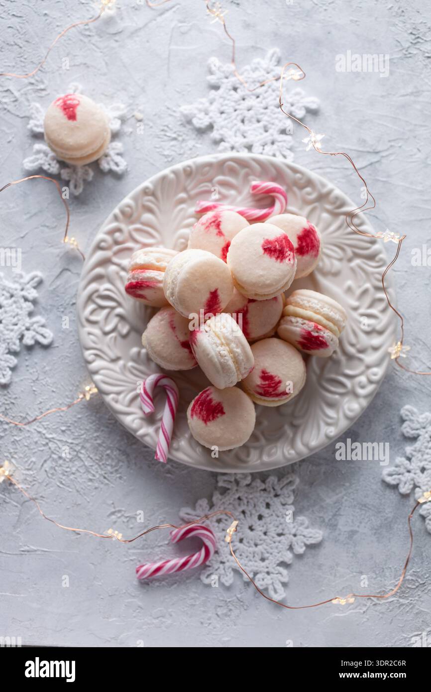 Photo de macarons blancs avec décoration rouge coup de pinceau disposés sur une assiette décorative entourée de cannes de bonbon crochet flocons de neige et lumière chaude d'étoile Banque D'Images