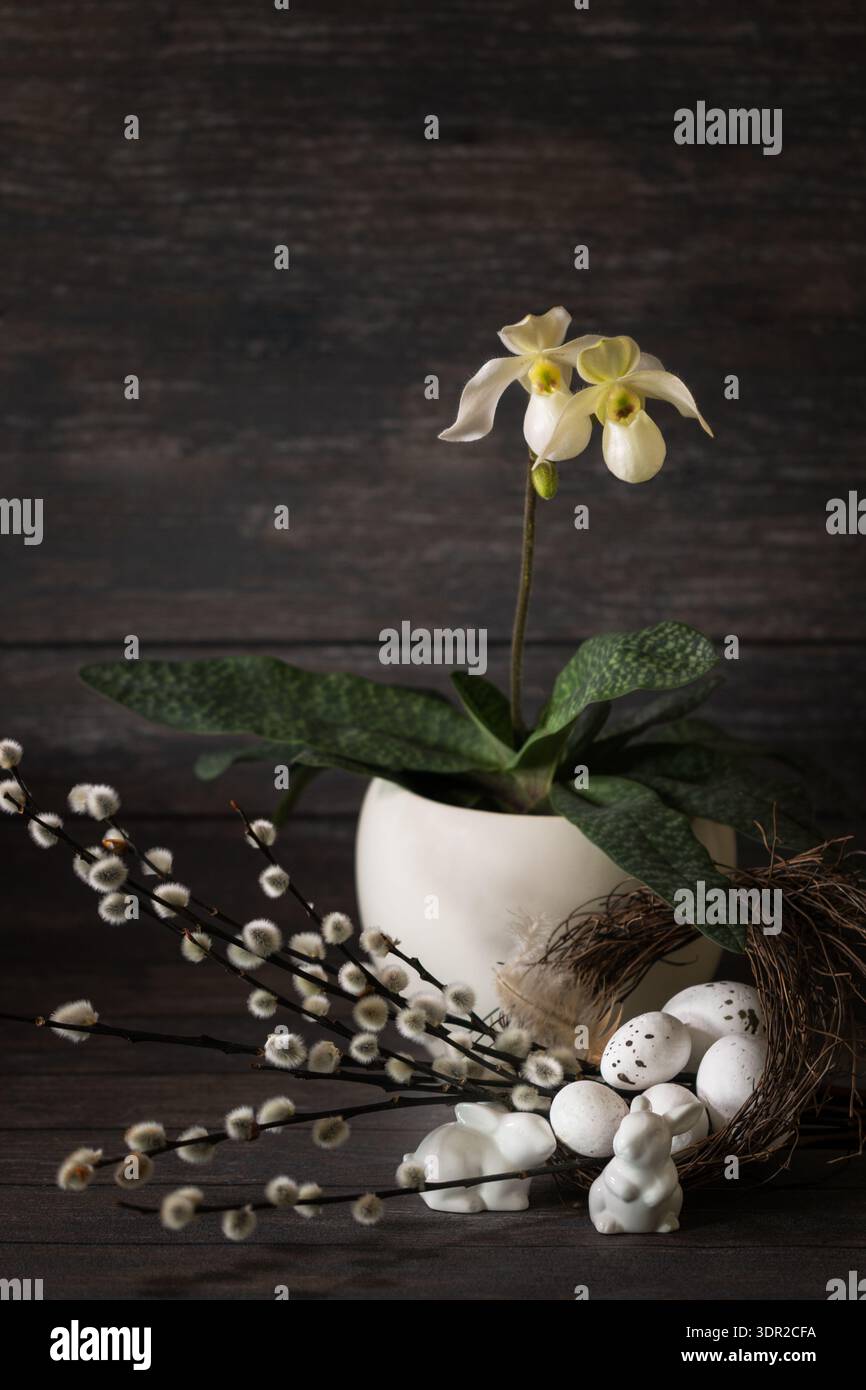 Joyeuses Pâques carte rustique avec fleur d'orchidée en pot. Une composition festive avec des œufs décoratifs, des brindilles de saule et des figures en céramique de lapins sur un sombre Banque D'Images