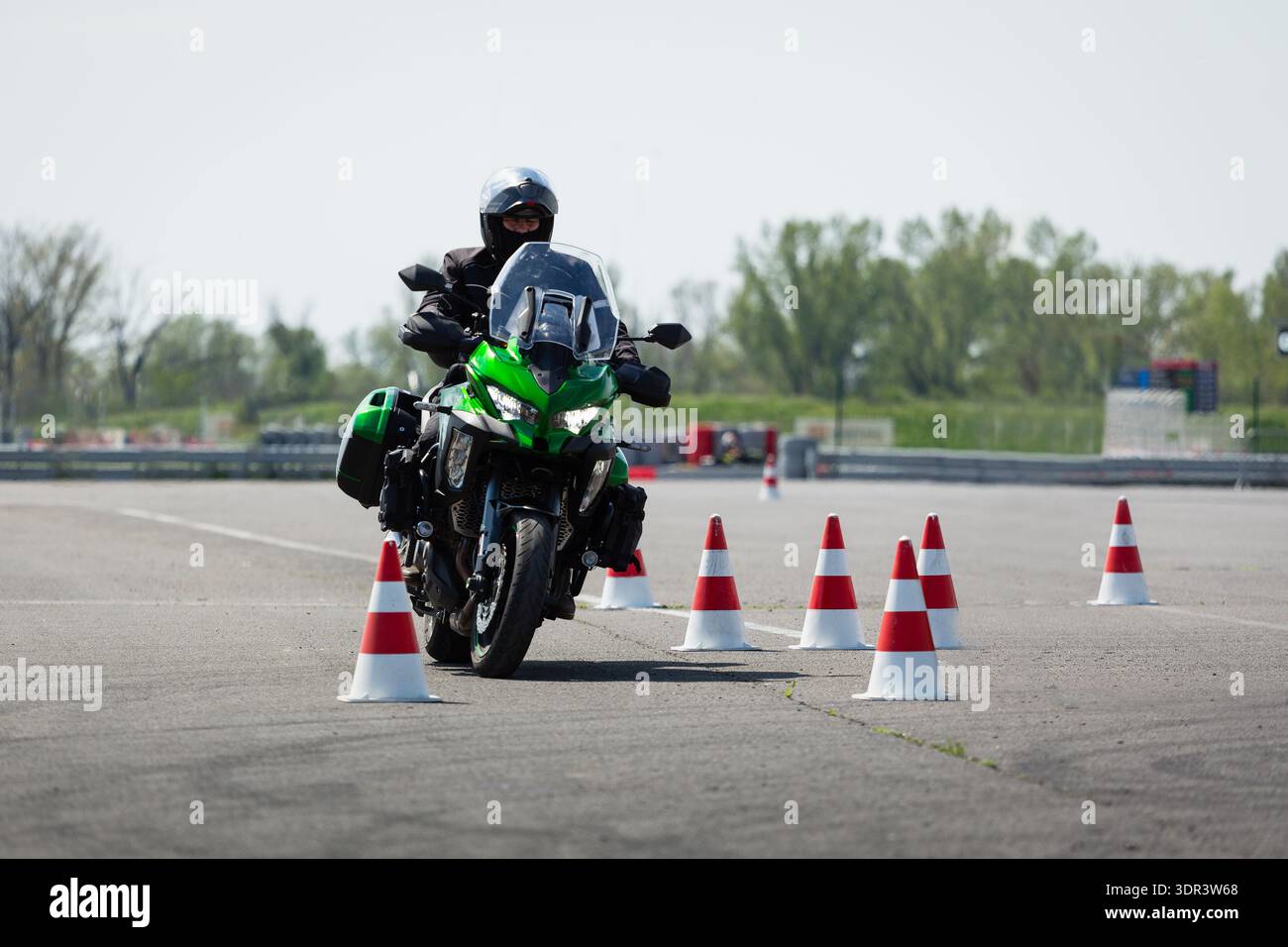 Un motocycliste sur une moto d'aventure verte naviguant à travers un parcours de slalom de cônes rouges et blancs sur un terrain d'entraînement pavé. Banque D'Images