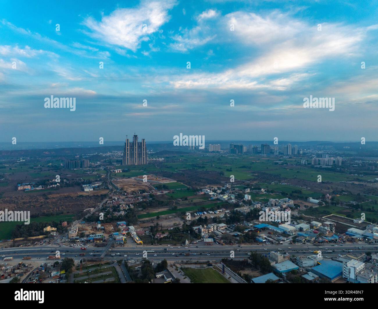 Vue aérienne des gratte-ciels modernes perce la ligne d'horizon au milieu d'une tapisserie de champs verdoyants et d'un étalement urbain dense, projetant de longues ombres sous un vaste ciel, Banque D'Images