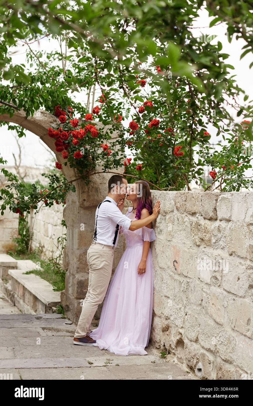 couple profite d'un moment ensemble dans un jardin, debout près d'un mur de pierre. Ils se penchent pour un baiser au milieu des fleurs en fleurs, capturées à la lumière du jour. Banque D'Images
