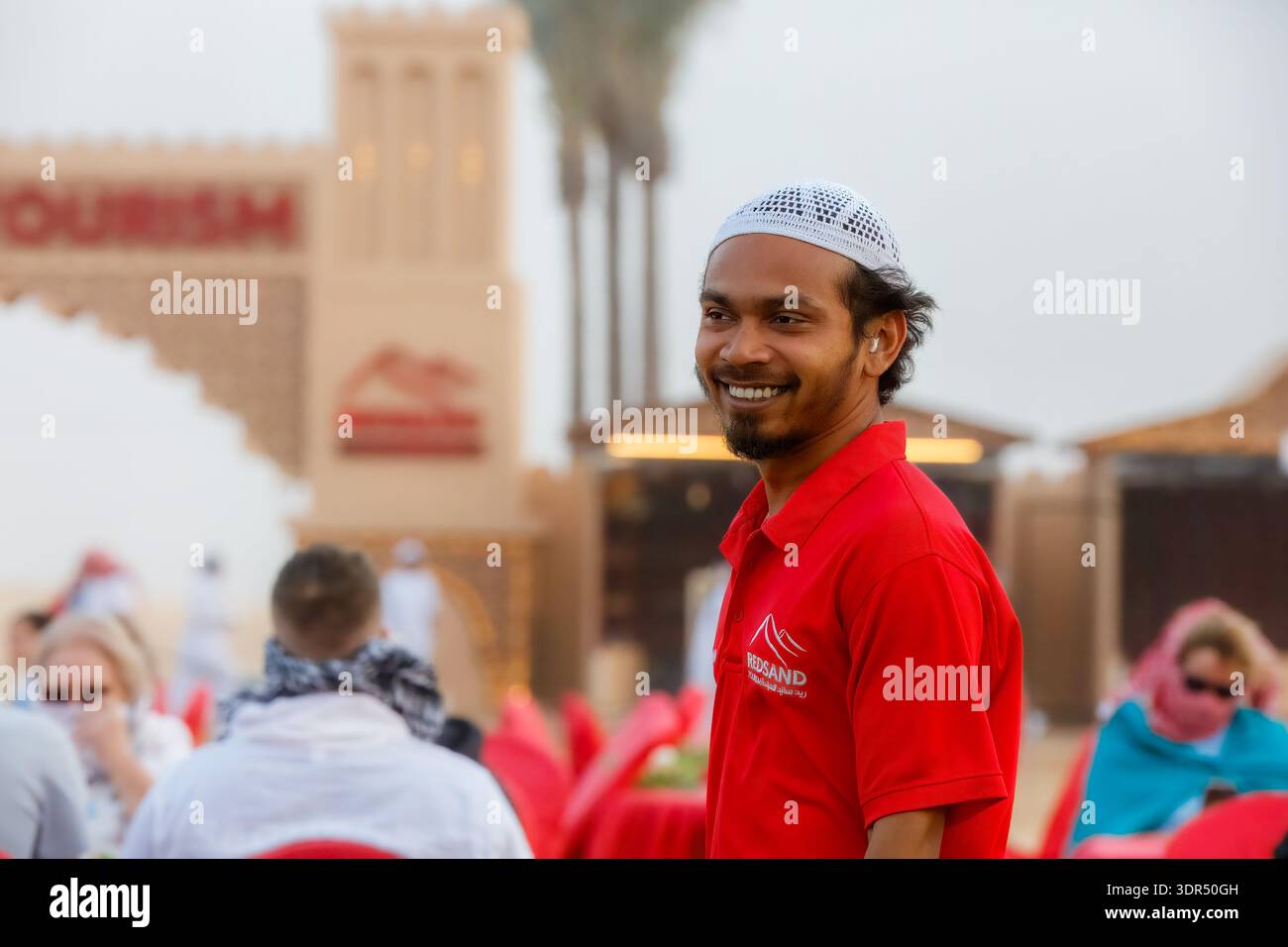 Dubaï, Émirats arabes Unis - 9 mars 2025 : un membre du personnel en chemise rouge et casquette blanche interagit avec les invités dans une salle à manger extérieure dans un tourisme dans le désert Banque D'Images