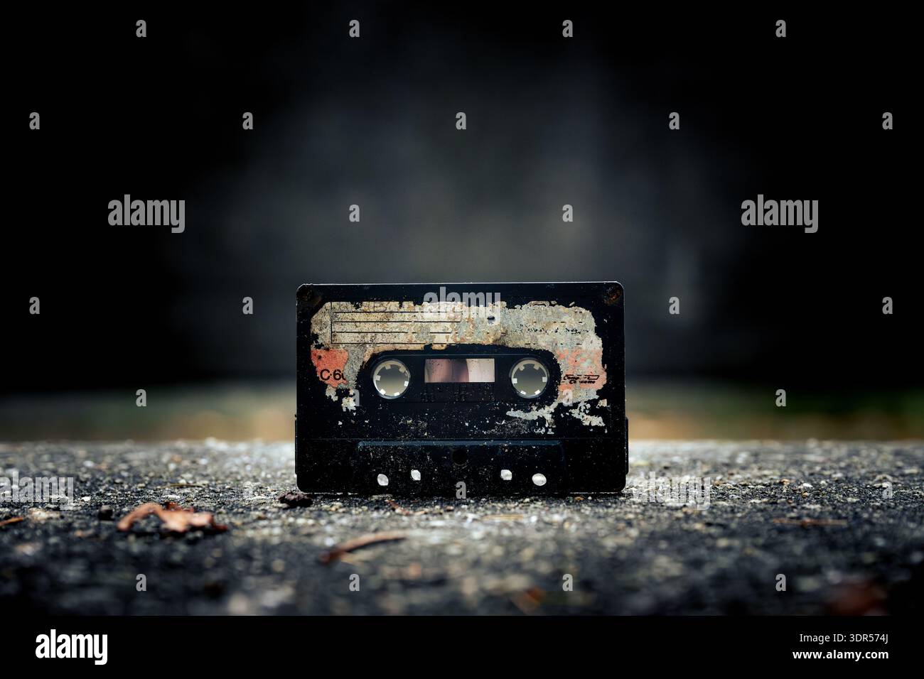 Une vieille cassette audio altérée est abandonnée sur une surface asphaltée sombre et texturée, montrant une usure importante et une étiquette qui se décolle. Banque D'Images