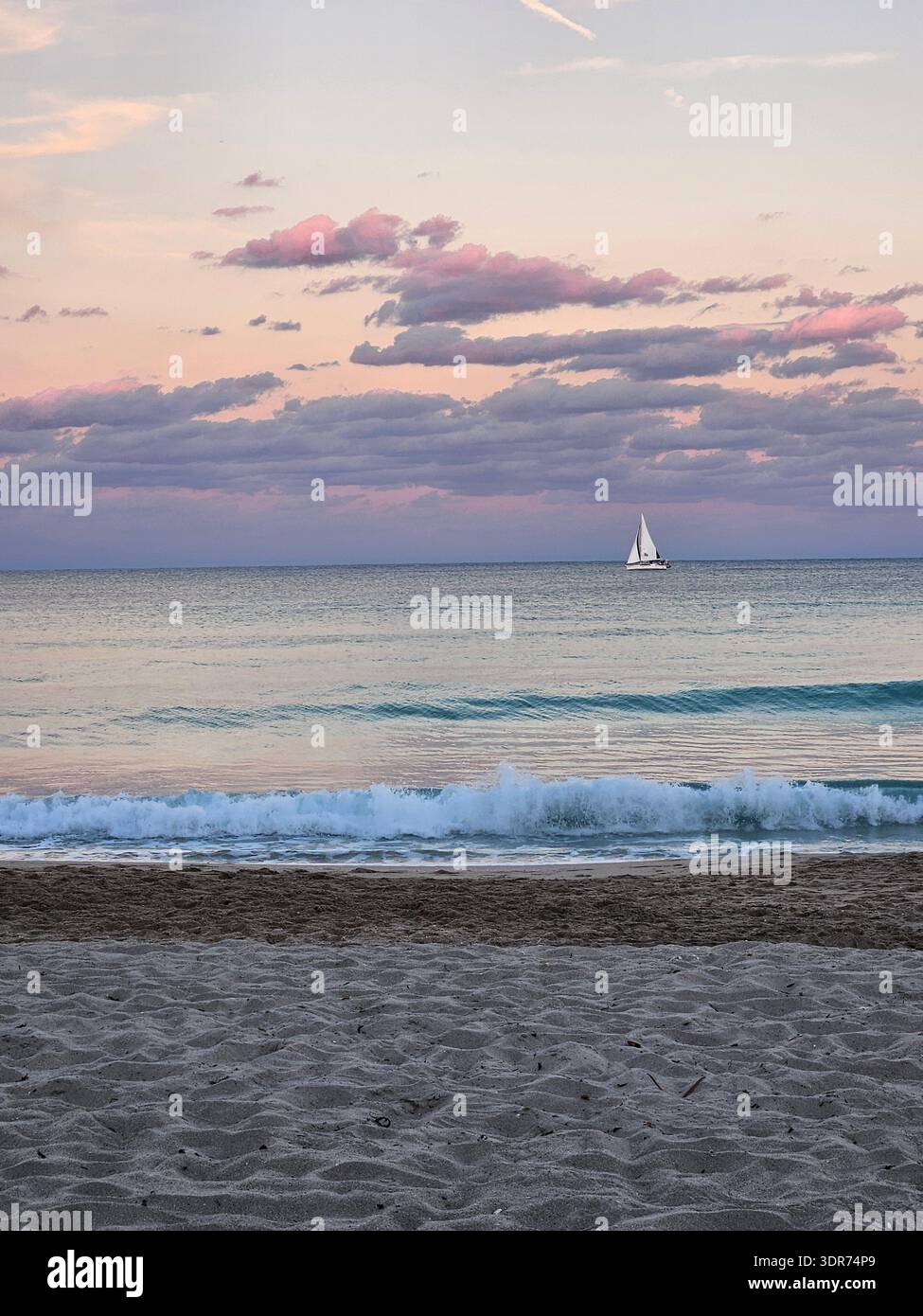 USAGE ÉDITORIAL EXCLUSIF - HOLLYWOOD BEACH FLORIDA : un voilier blanc solitaire glisse paisiblement sur le calme océan Atlantique à Hollywood Beach, Floride, pendant un magnifique coucher de soleil pastel. Le ciel est rempli de nuages stratocumulus en couches dans des teintes de lavande douce, rose foncé et violet sourd, créant une scène nautique tranquille et romantique. Le premier plan présente le sable texturé de la plage, menant le regard vers le doux surf turquoise et l'horizon lointain où le ciel rencontre la mer dans un affichage serein de la beauté naturelle du sud de la Floride. - Image de stock capturée avec un smartphone
