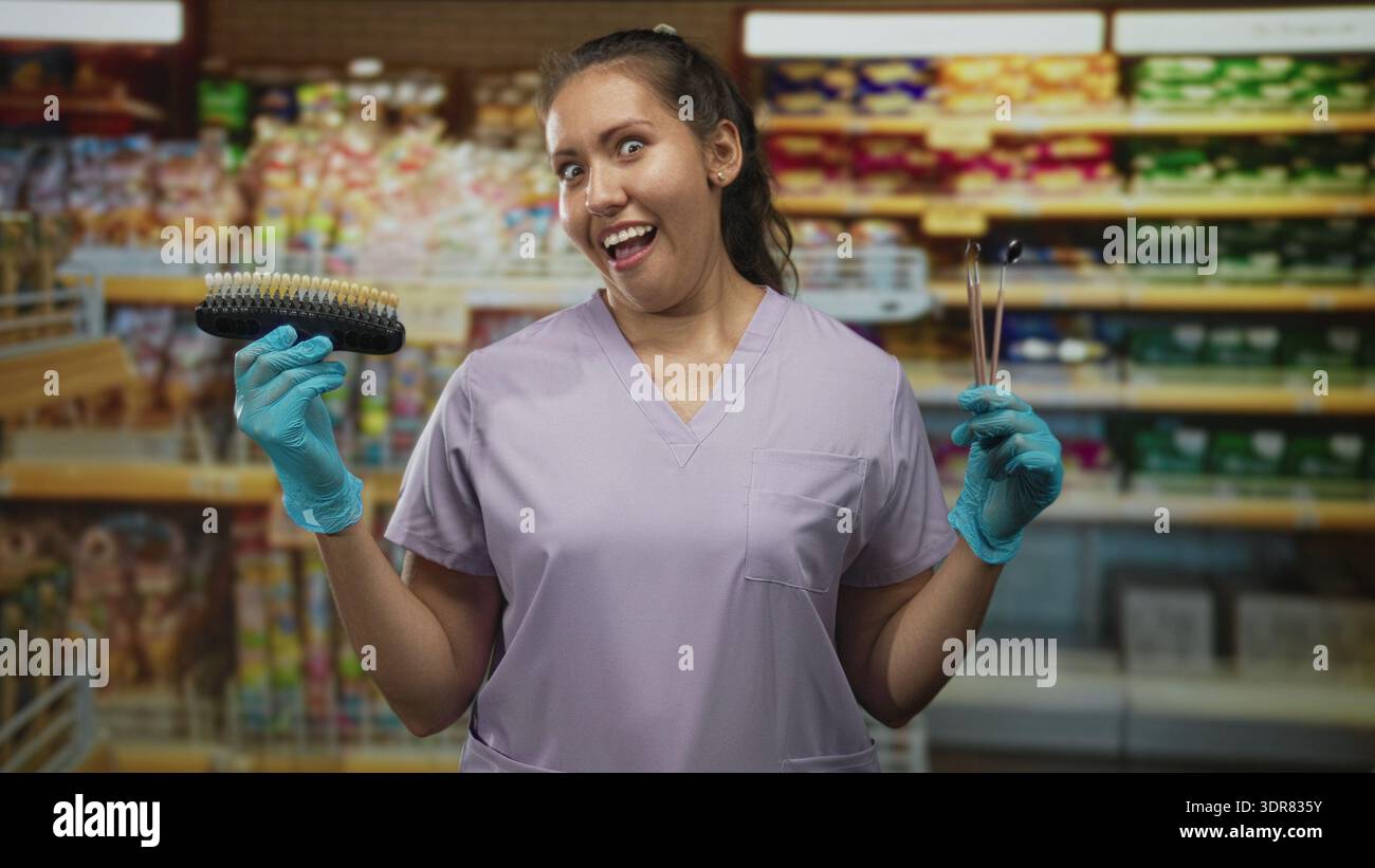 Femme dentiste tient guide de teinte de dent et sondes dentaires, souriant avec les mains gantées dans la construction de supermarché ; confiance ludique. Banque D'Images