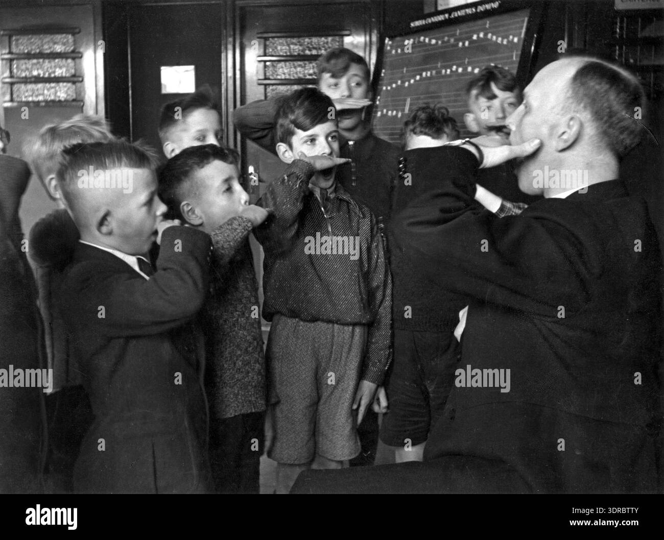 La photographie montre une leçon de chant du chœur de garçons de la cathédrale à la basilique catholique romaine de Bavo à Haarlem en 1941. Le professeur de musique, Dr de Kat, démontre la formation vocale avec sa main tandis que les garçons l'imitent. L’image documente l’éducation chorale néerlandaise, la musique religieuse, les pratiques chorale des garçons, l’instruction vocale, les traditions liturgiques, et pédagogie musicale du début du xxe siècle. Banque D'Images