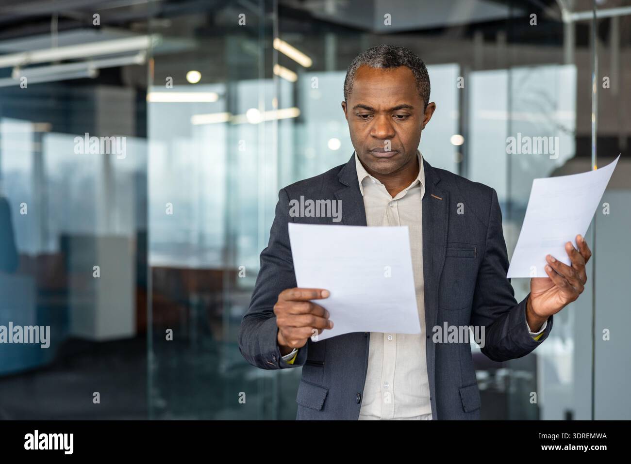 Homme d'affaires afro-américain sérieux debout dans un bureau moderne, se concentrant tout en examinant plusieurs documents commerciaux et en comparant les informations pour la prise de décision critique Banque D'Images