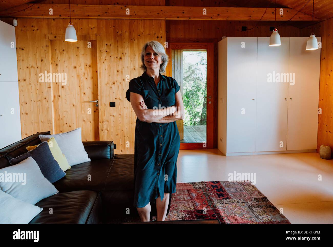 Femme senior élégante avec les cheveux gris souriant avec confiance, les bras croisés tout en se tenant debout dans un salon lumineux et moderne avec des murs de pin et un canapé confortable Banque D'Images