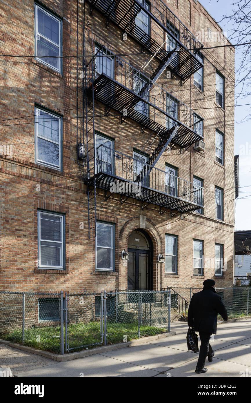 Un homme vêtu de noir marche le long d'un trottoir devant un immeuble d'appartements en brique à plusieurs étages avec des évasions de feu en fer à Jersey City, NJ Banque D'Images