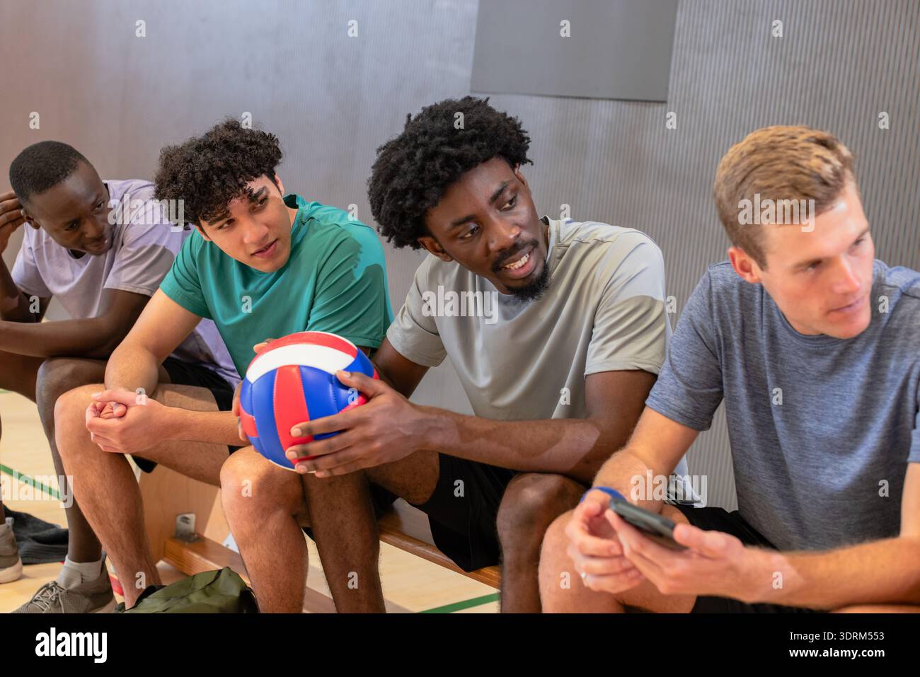 Divers coéquipiers masculins de volley-ball se reposant sur un banc en bois dans la salle de gym tenant le volley-ball et le smartphone. Athlètes, travail d'équipe, occasionnel, Banque D'Images