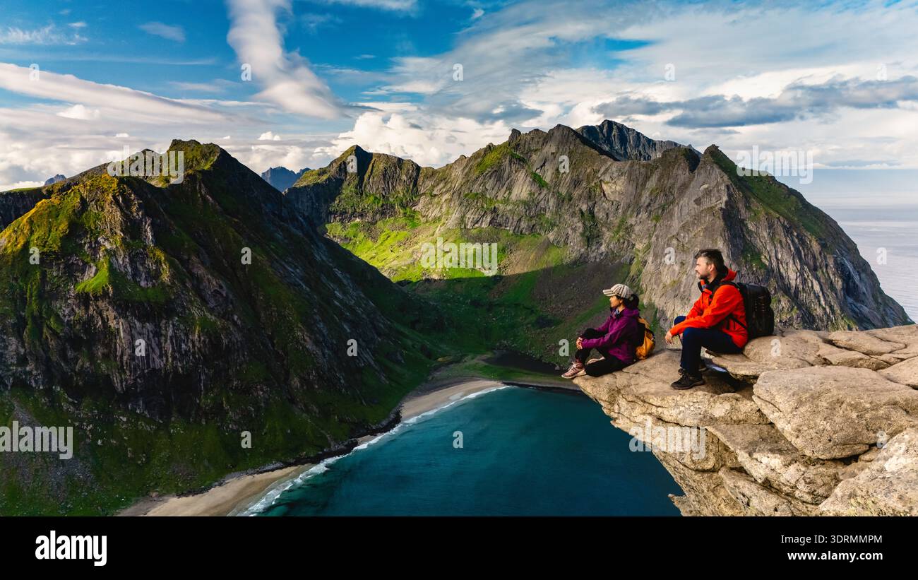 Les aventuriers se détendent sur une corniche rocheuse, en profitant d'une vue imprenable sur la plage de Ryten Kvalvika et les montagnes majestueuses. Verdure luxuriante et dr. Banque D'Images