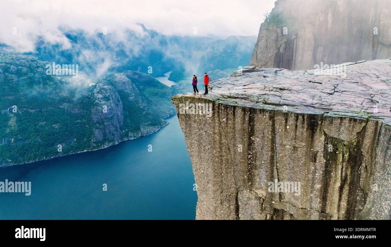 Deux randonneurs se tiennent au bord de la falaise Preikestolen, surplombant un magnifique fjord en Norvège. La brume matinale recouvre doucement le Banque D'Images