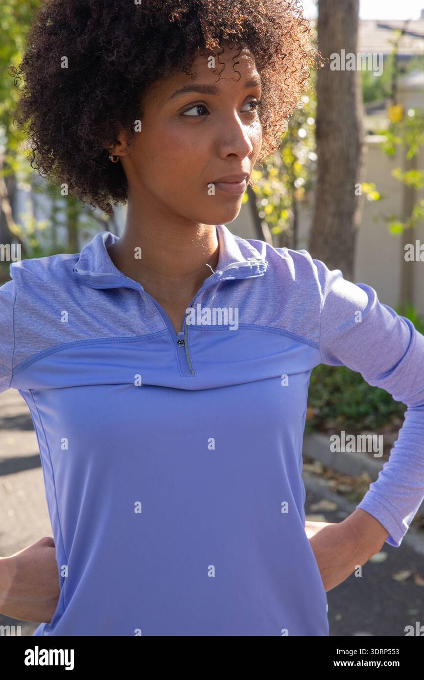 Femme afro-américaine debout sur la rue résidentielle dans le quart-zip de lavande haut, regardant à droite. Vêtements de sport, décontracté, vibrant, ou Banque D'Images