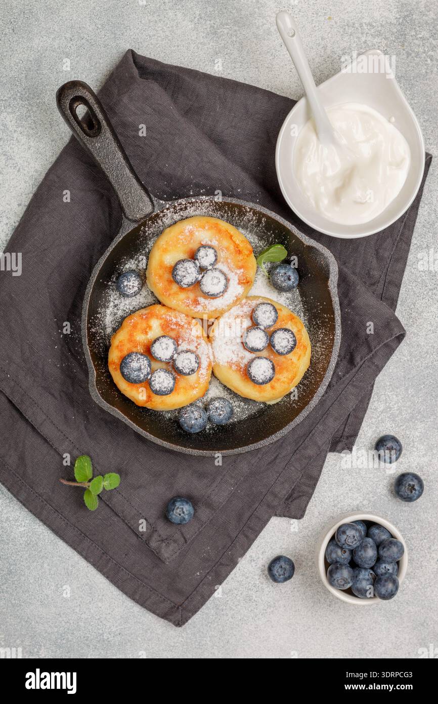 Crêpes au fromage cottage, syrniki, beignets de lait caillé avec baies fraîches, myrtille, sucre en poudre et crème sure dans une poêle en fonte. Banque D'Images