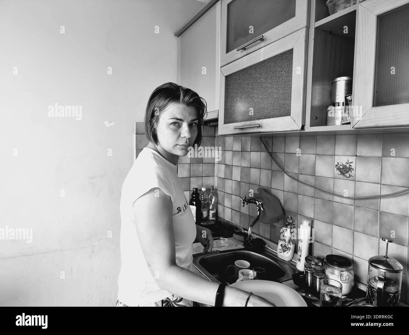 Femme debout dans une cuisine à la maison laver la vaisselle dans l'évier, effectuer les tâches ménagères quotidiennes et maintenir la maison dans un présentat noir et blanc Banque D'Images