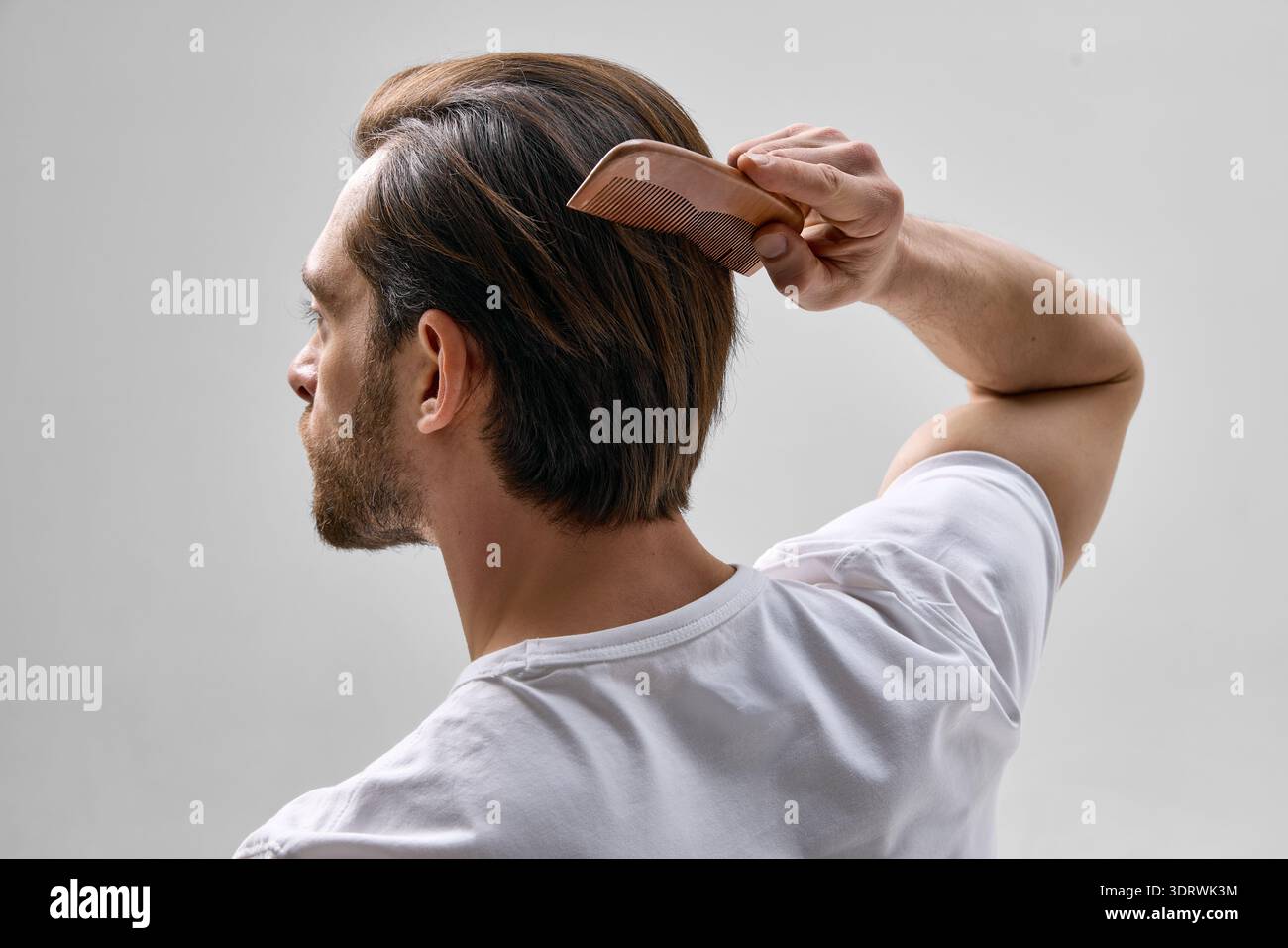Vue arrière de l'homme dans la trentaine avec la barbe courte brossant les cheveux avec peigne en bois sur fond clair, portant t-shirt blanc dans un cadre minimal. Banque D'Images