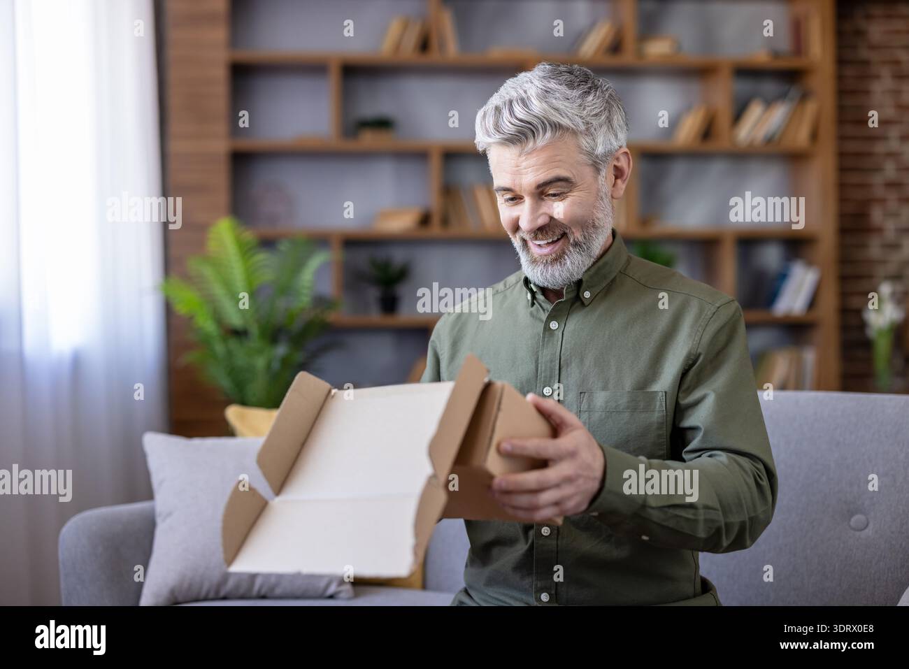 Homme senior avec les cheveux gris et la barbe assis sur un canapé, ouvrant un colis de carton à la maison, éprouvant la joie et la satisfaction de son nouvel achat ou cadeau d'une commande en ligne Banque D'Images