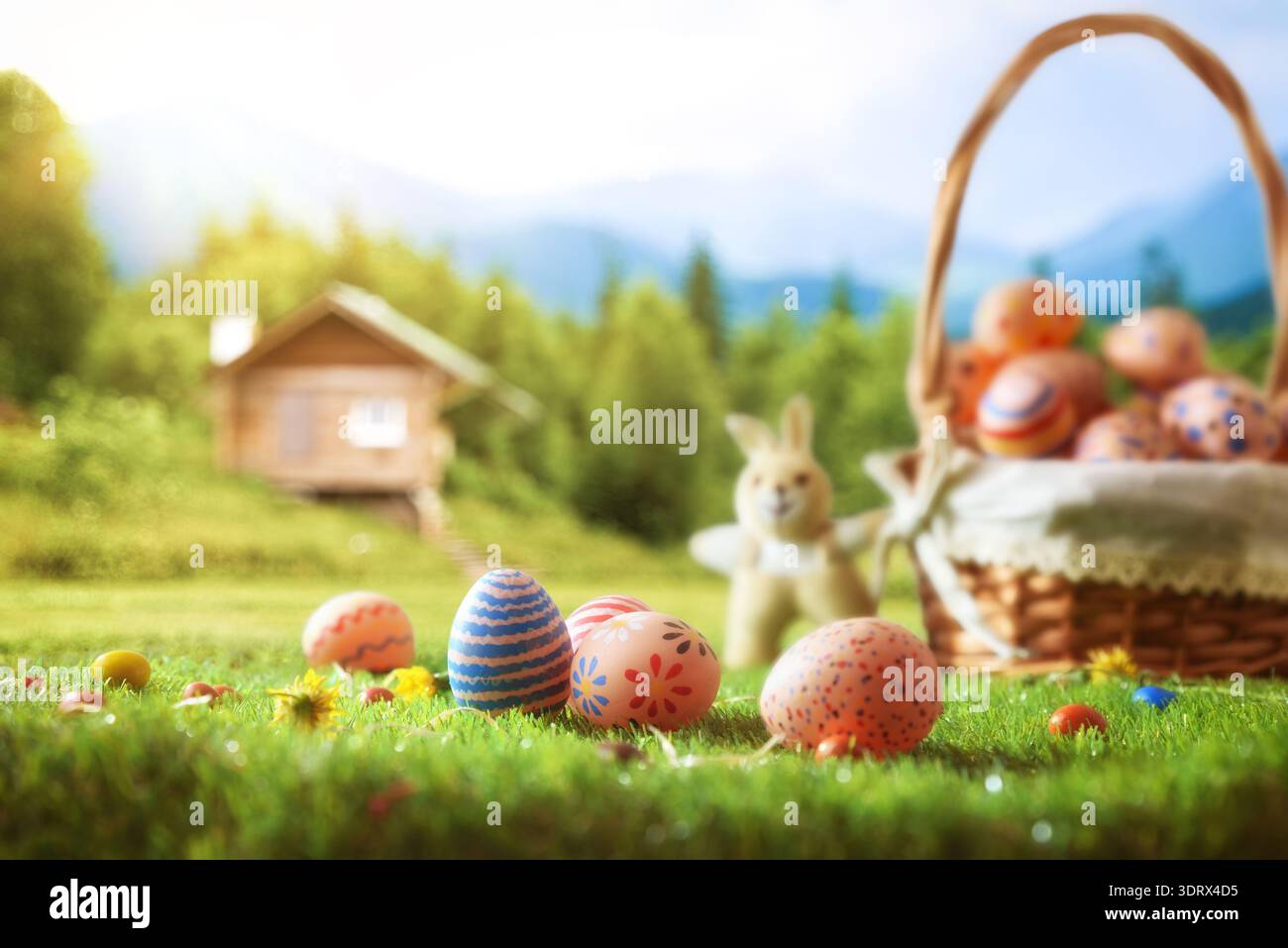 Oeufs de Pâques décorés dans un panier sur l'herbe verte avec un petit jouet lapin. Paysage printanier ensoleillé avec cabine en bois et montagnes floues en arrière-plan. Banque D'Images