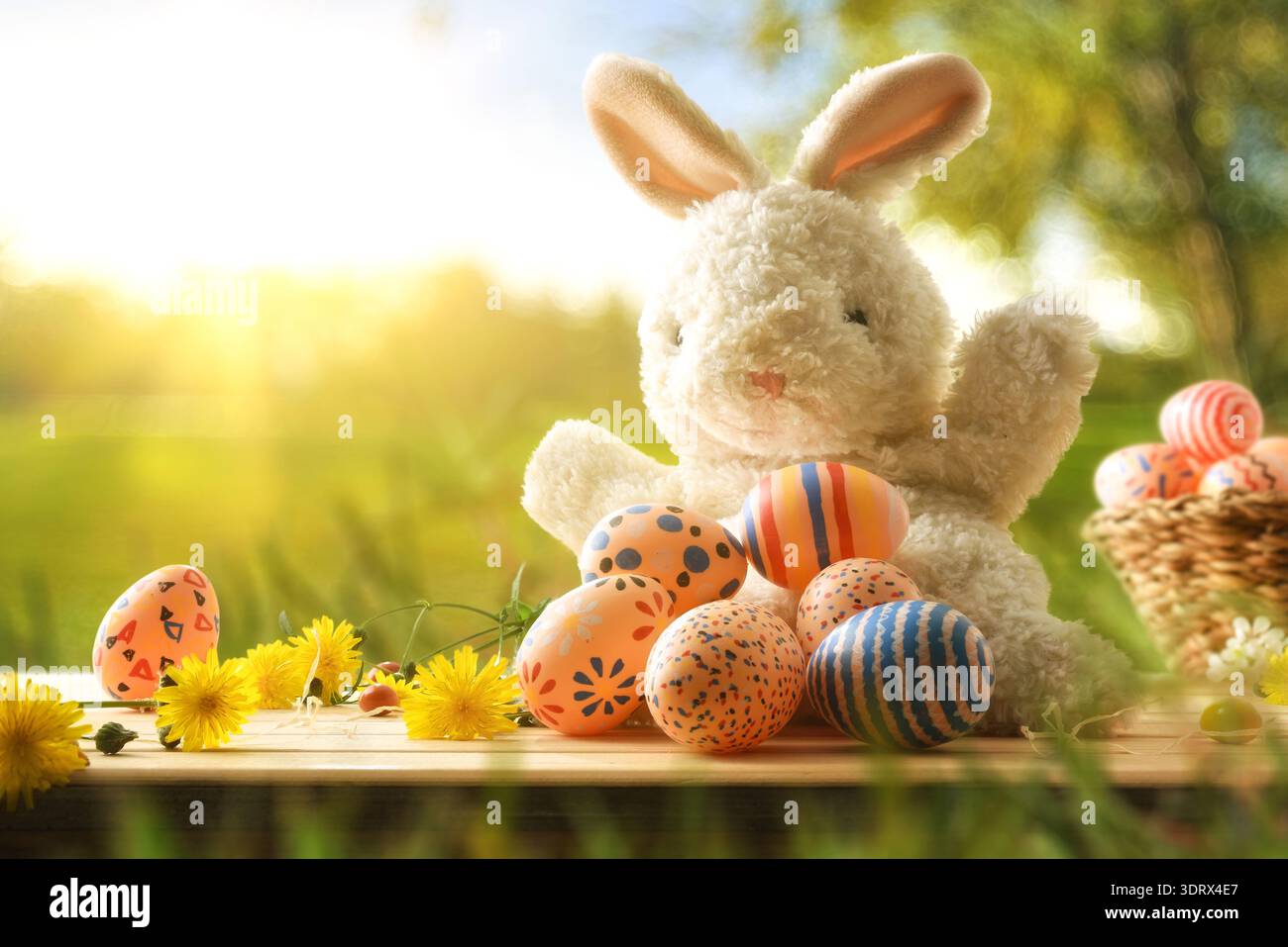 Lapin jouet moelleux avec des œufs de Pâques peints et des fleurs jaunes sur une table en bois, fond de jardin de printemps ensoleillé avec un bokeh doux et une lumière chaude. Banque D'Images