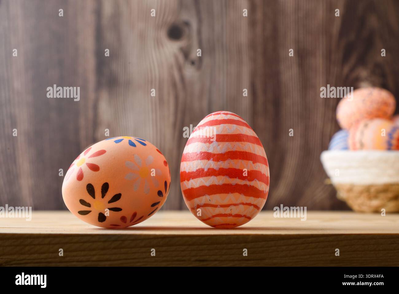 Deux oeufs de Pâques peints à la main avec des motifs floraux et rayés sur une étagère en bois, avec un panier d'oeufs flou en arrière-plan. Banque D'Images