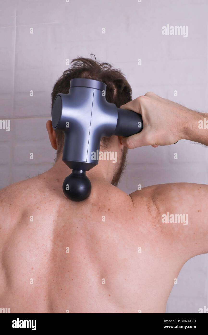 Gros plan arrière de l'homme appliquant un pistolet de massage à la région supérieure du cou et des épaules, stimulation musculaire profonde après l'entraînement Banque D'Images
