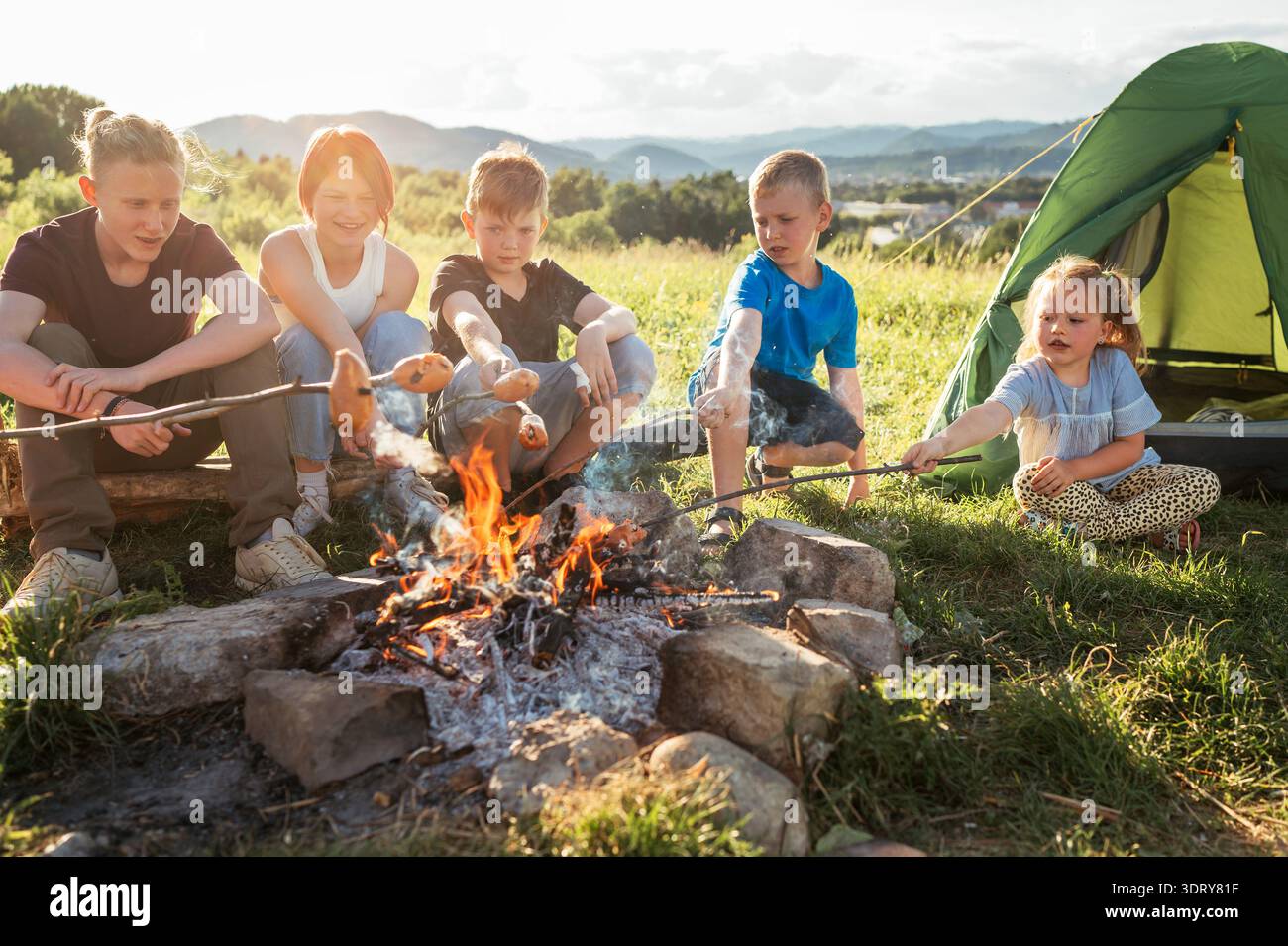 Groupe de cinq enfants garçons et filles riaient joyeusement et rôtissaient des saucisses sur des bâtons au-dessus d'une flamme de feu de camp près de la tente verte. Temps actif en plein air spendi Banque D'Images