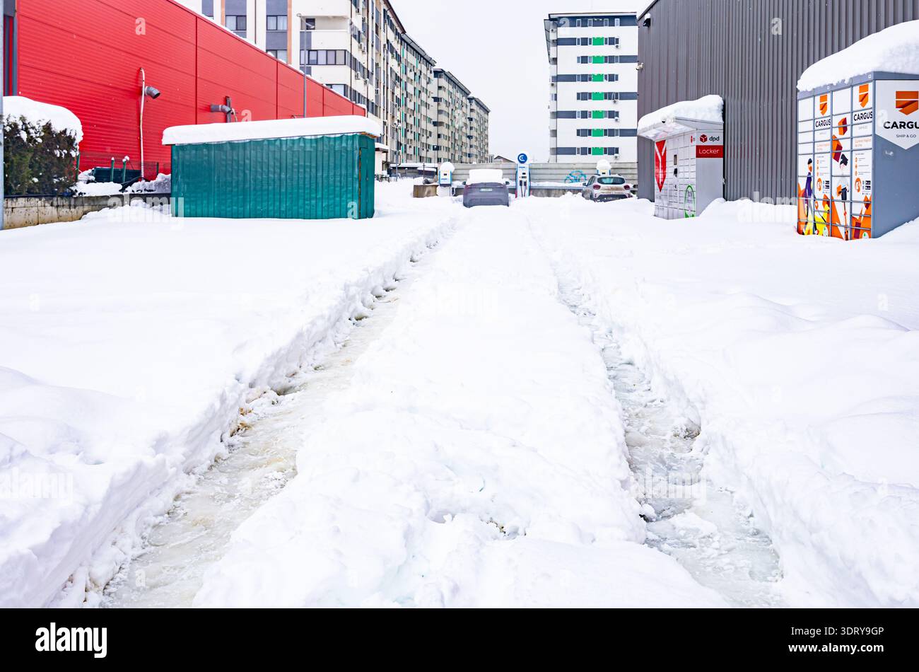 Rue de ville d'hiver avec de profondes traces de pneus dans la neige épaisse menant à des casiers à colis en libre-service et bâtiment résidentiel Banque D'Images