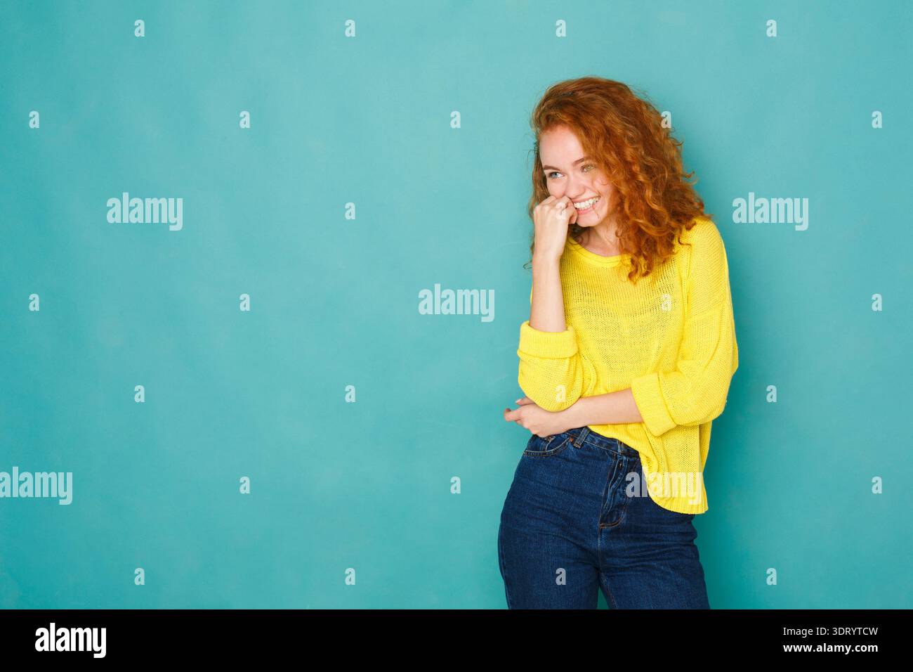 Femme souriante tout en se tenant debout contre un mur bleu avec une pose ludique et portant un pull jaune Banque D'Images