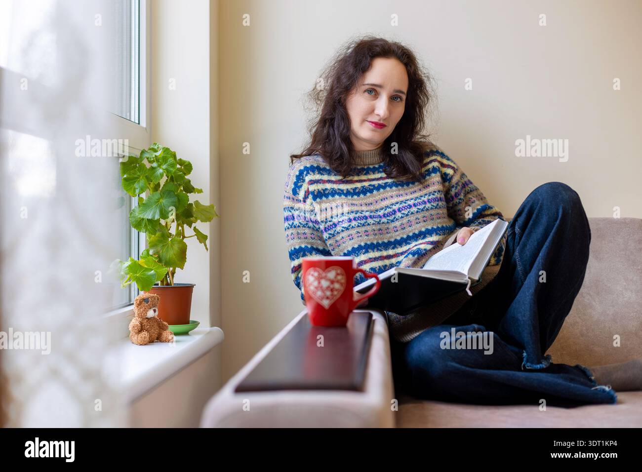 Femme d'âge moyen se relaxant avec un livre et une tasse. Portrait confortable de style de vie d'une femme brune d'âge moyen assise sur un canapé près de la fenêtre, lisant un livre Banque D'Images
