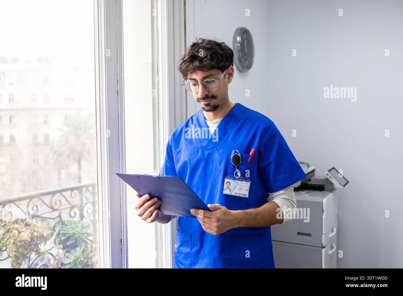 Un professionnel de la santé en gommages bleus examine des documents médicaux dans un bureau lumineux. La scène reflète un environnement de soins de santé moderne, mettant l'accent sur Tho Banque D'Images