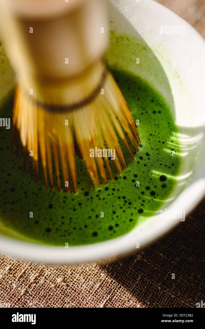 Un fouet traditionnel en bambou remue délicatement le thé vert matcha vif et mousseux dans un bol blanc. La scène capture l'essence du rituel japonais de fabrication du thé Banque D'Images