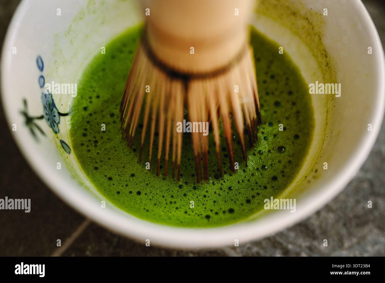 Gros plan d'un fouet en bambou remuant du matcha vert éclatant dans un bol en céramique. Le processus de mélange crée une texture lisse et mousseuse, mettant en évidence t Banque D'Images