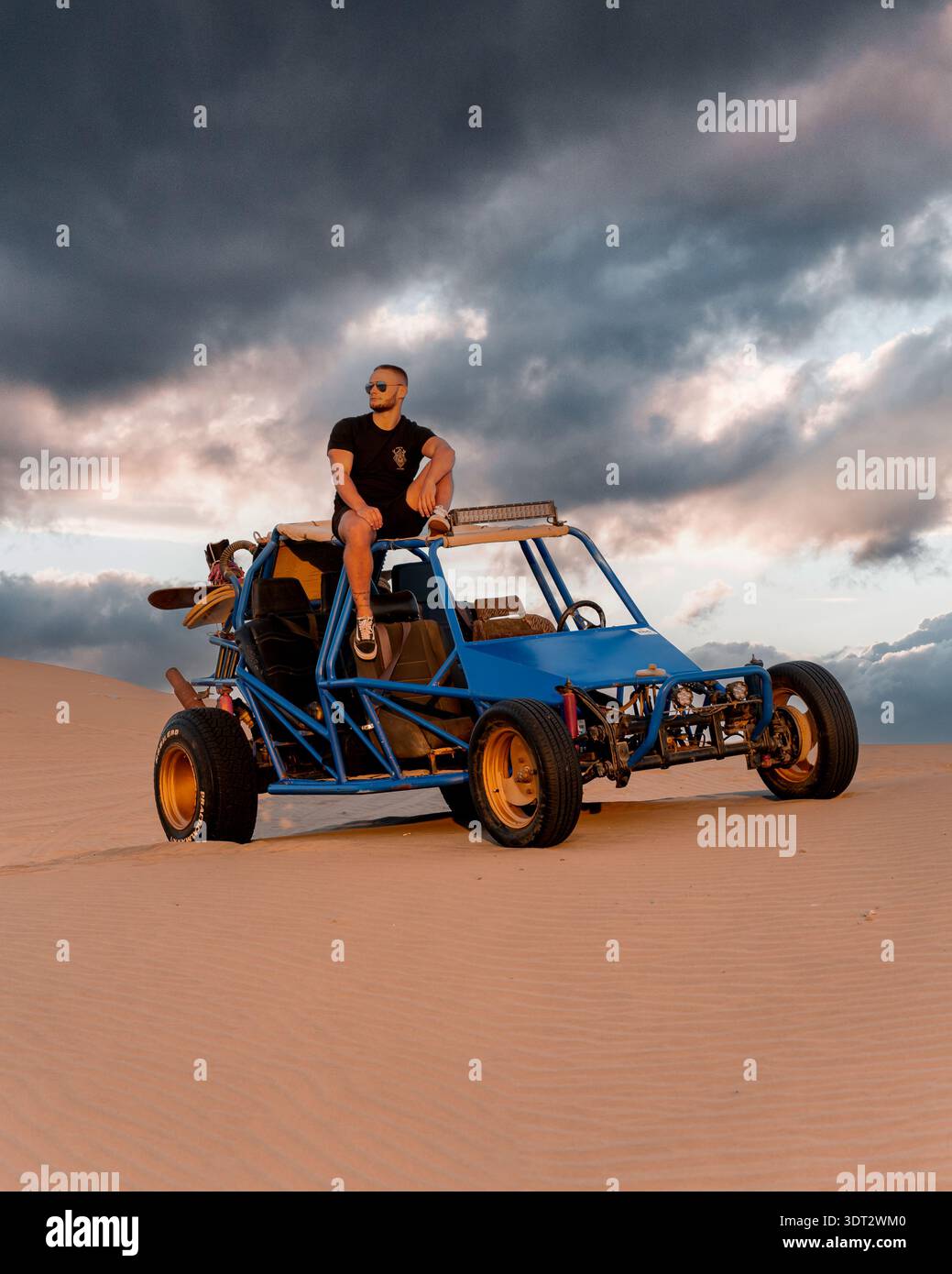 Vue d'un homme perché au sommet d'un buggy dunaire bleu avec des roues dorées sur la toile de fond d'un paysage sablonneux et d'un ciel dramatique, ICA, ICA, Pérou. Banque D'Images