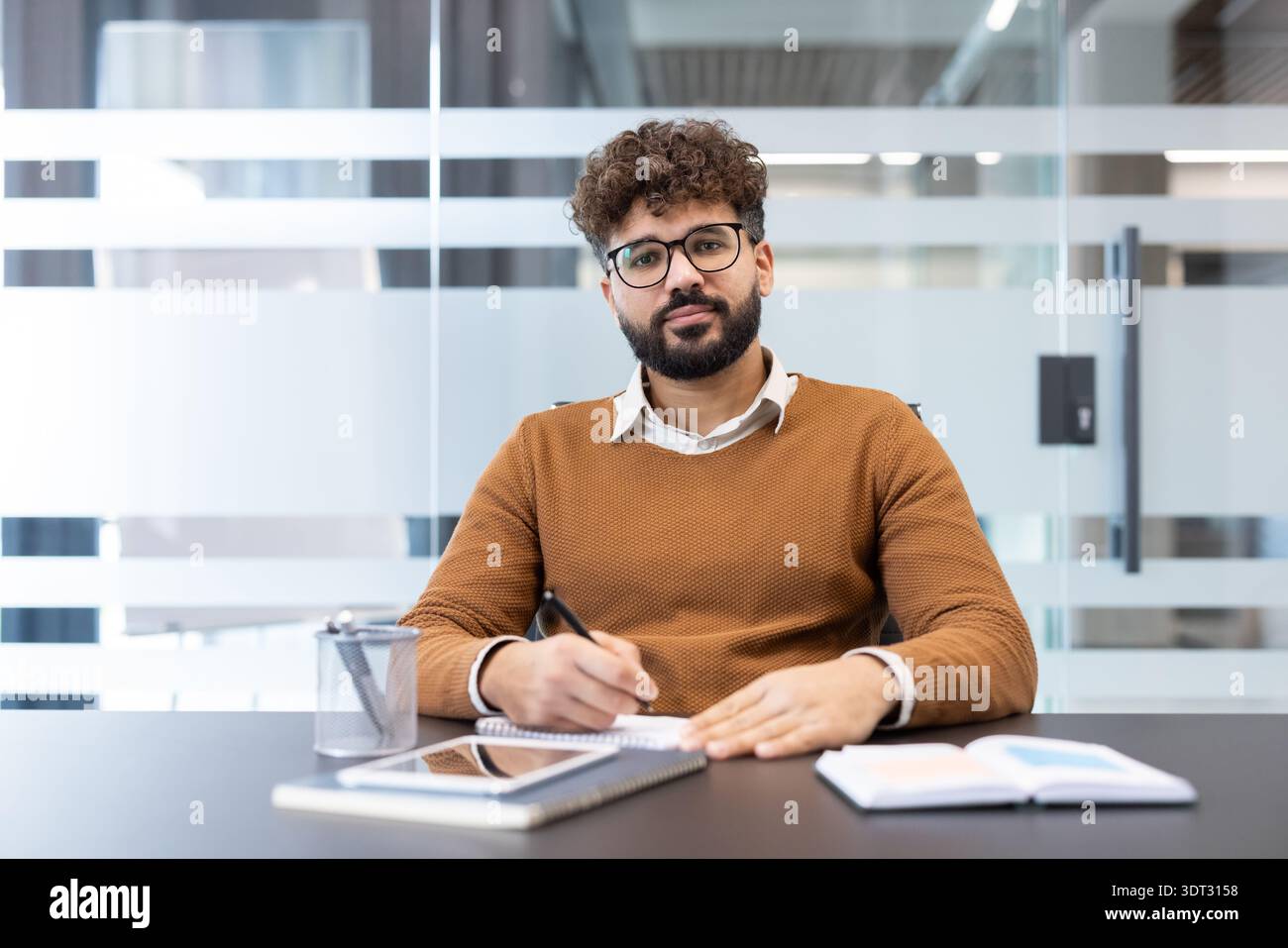 Jeune homme d'affaires avec des vêtements décontractés et des lunettes assis au bureau, tenant un stylo et écrivant dans un cahier, se concentrant sur la paperasse et Banque D'Images