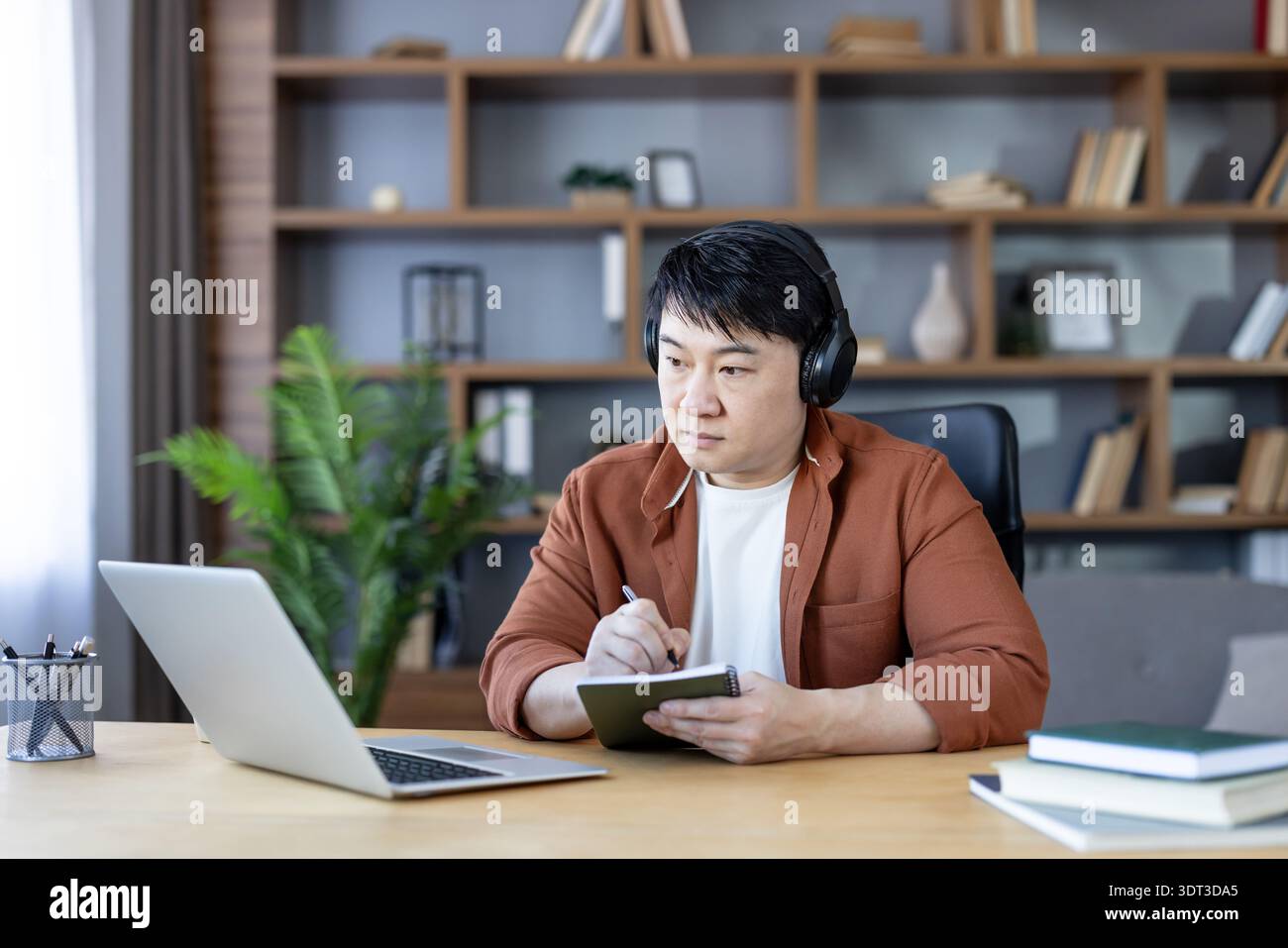 Homme asiatique dans le casque apprenant intentionnellement de son ordinateur portable, prenant des notes dans un ordinateur portable tout en participant à une éducation à distance ou une réunion virtuelle depuis son bureau confortable à domicile Banque D'Images