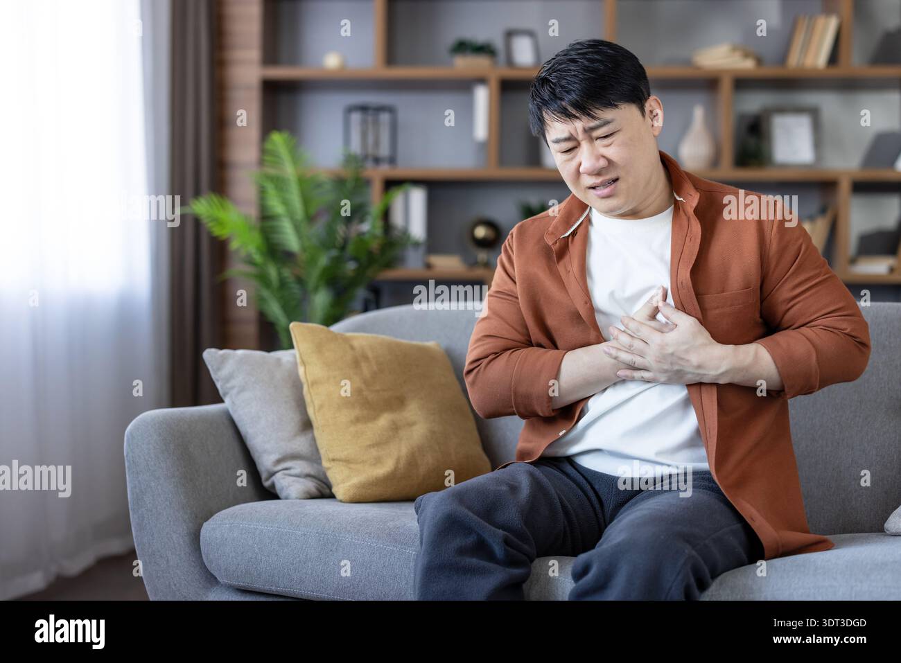 Homme asiatique éprouvant une douleur thoracique sévère et un inconfort d'une crise cardiaque, assis sur un canapé à la maison et serrant d'urgence sa poitrine avec les deux mains Banque D'Images