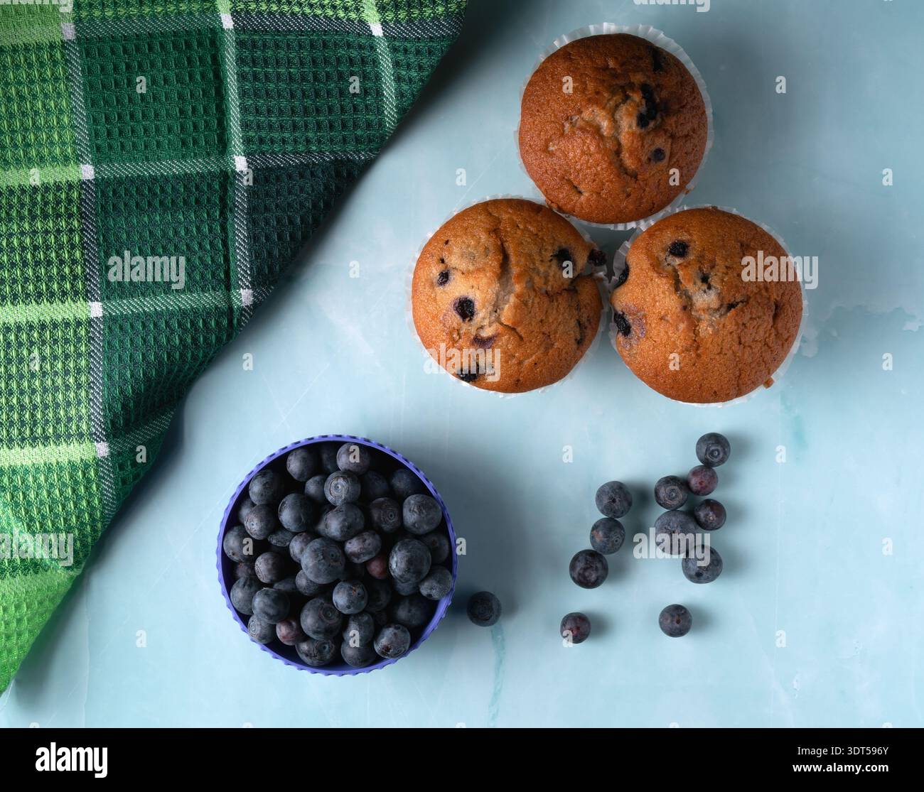 Vue aérienne de trois muffins aux myrtilles dorées servis avec un bol de myrtilles biologiques fraîches sur un fond esthétique lumineux Banque D'Images