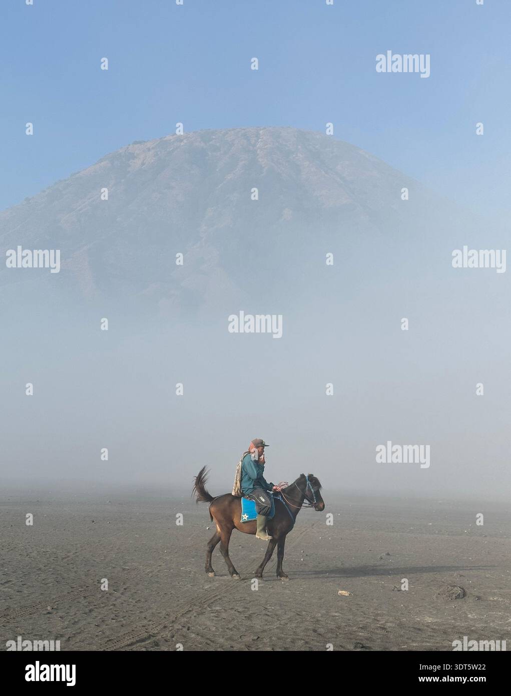 Un cavalier seul à cheval traverse une plaine volcanique pâle sous une silhouette montagneuse brumeuse - Mount bromo Cowboy Banque D'Images